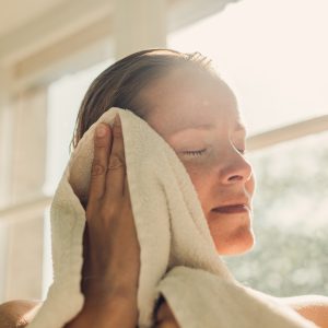Close-up of a person gently drying the side of their face with a white towel near a sunlit window, with soft natural light illuminating the scene.