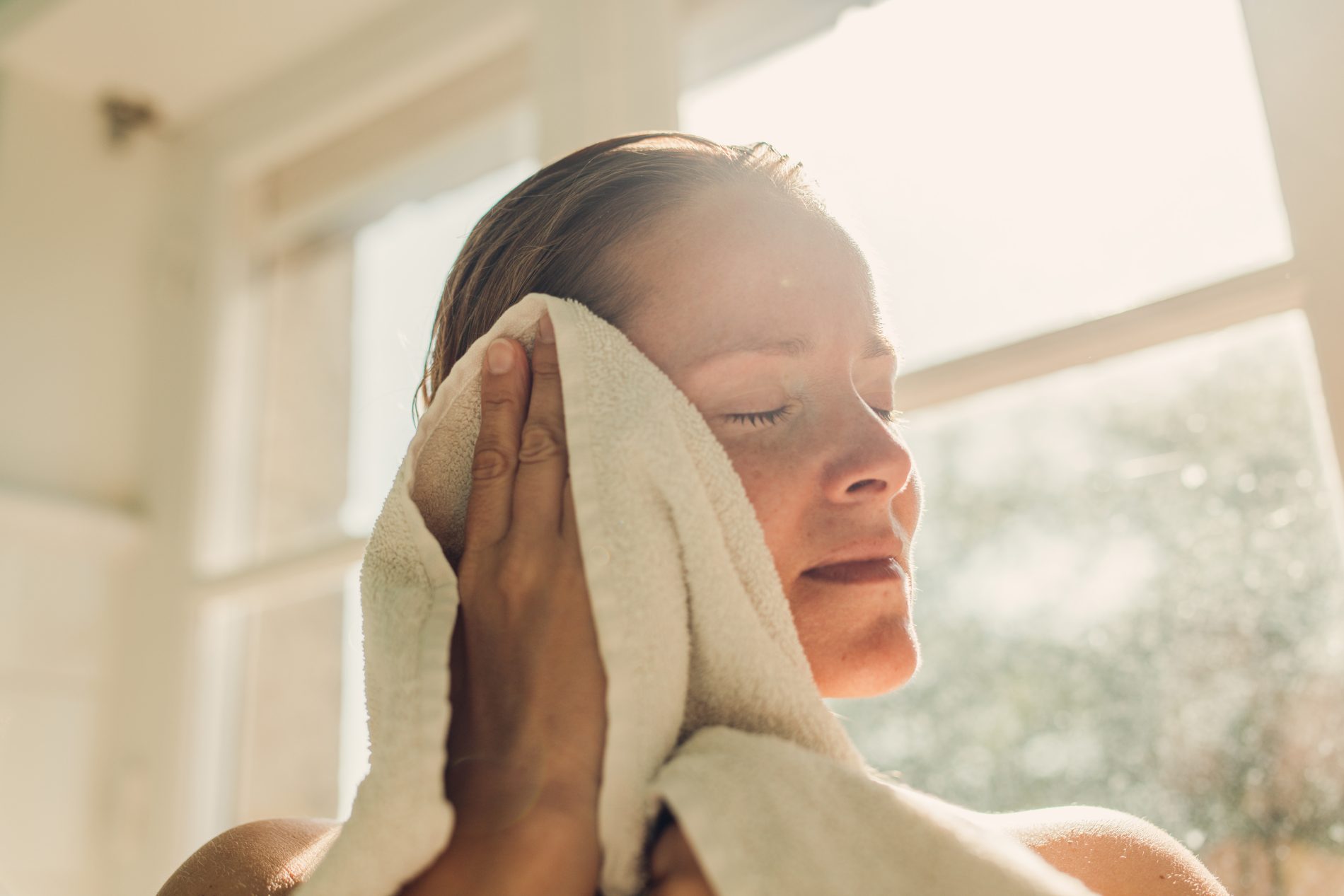 Close-up of a person gently drying the side of their face with a white towel near a sunlit window, with soft natural light illuminating the scene.